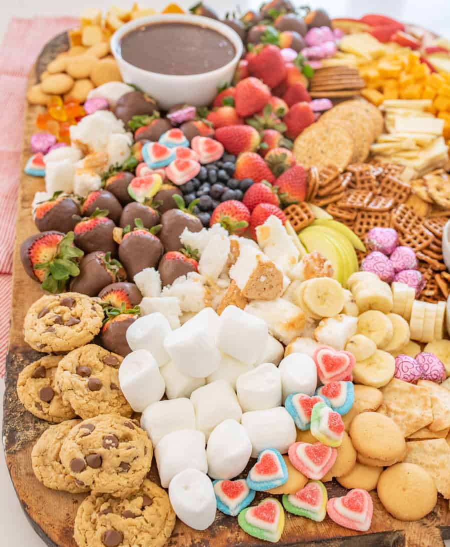 A large wooden board covered in cheeses, crackers, jam, mustard, toothpicks, chocolate fondue and things to dip in the fondue including fruit, chocolate chip cookies, vanilla wafers, banana slices, marshmallows, and garnished with heart-shaped gum drops and chocolates.