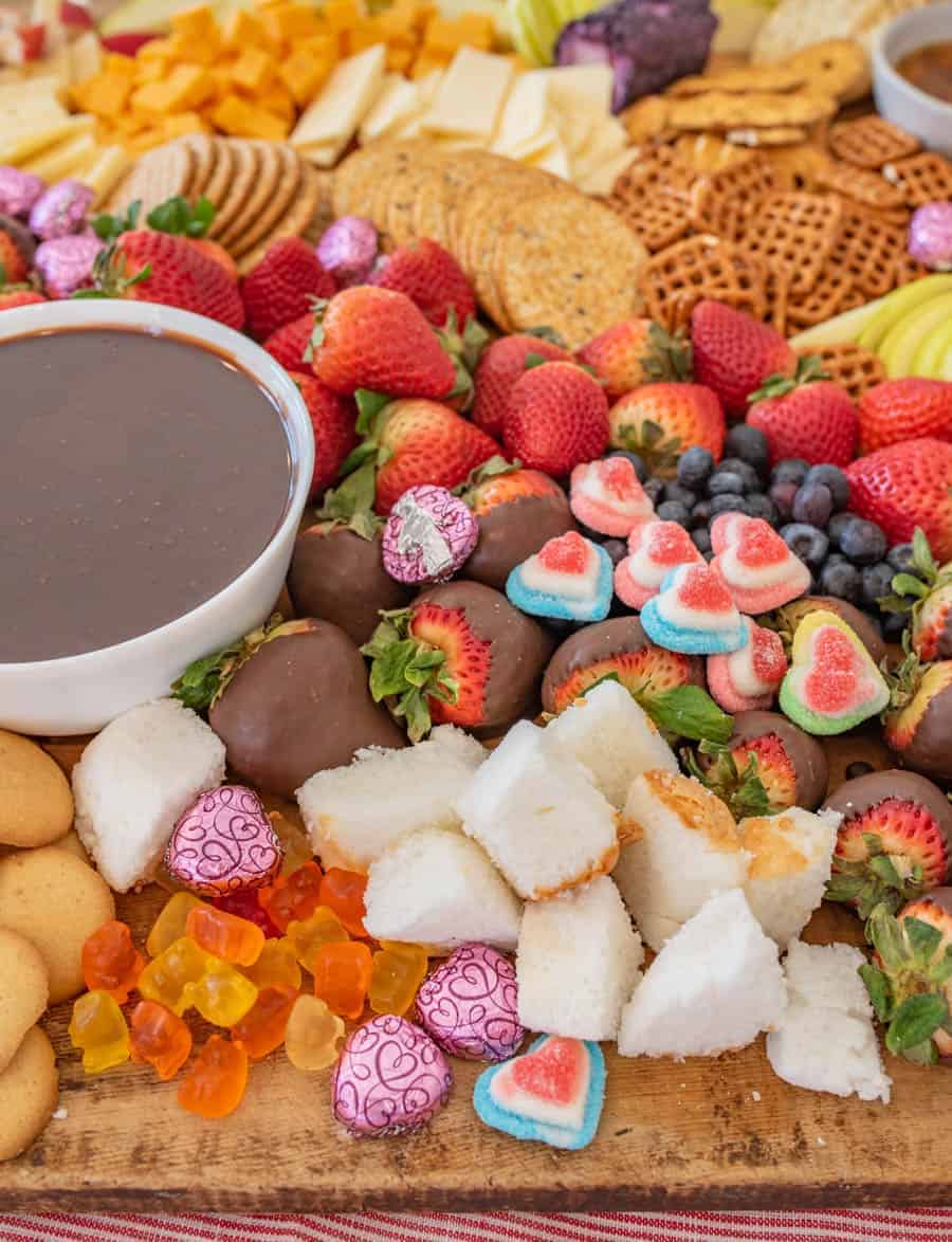 A close up of the chocolate fondue on the cheese board with things to dip in the fondue including pretzels, apple slices, strawberries, blueberries, and garnished with heart-shaped chocolates and gum drops.