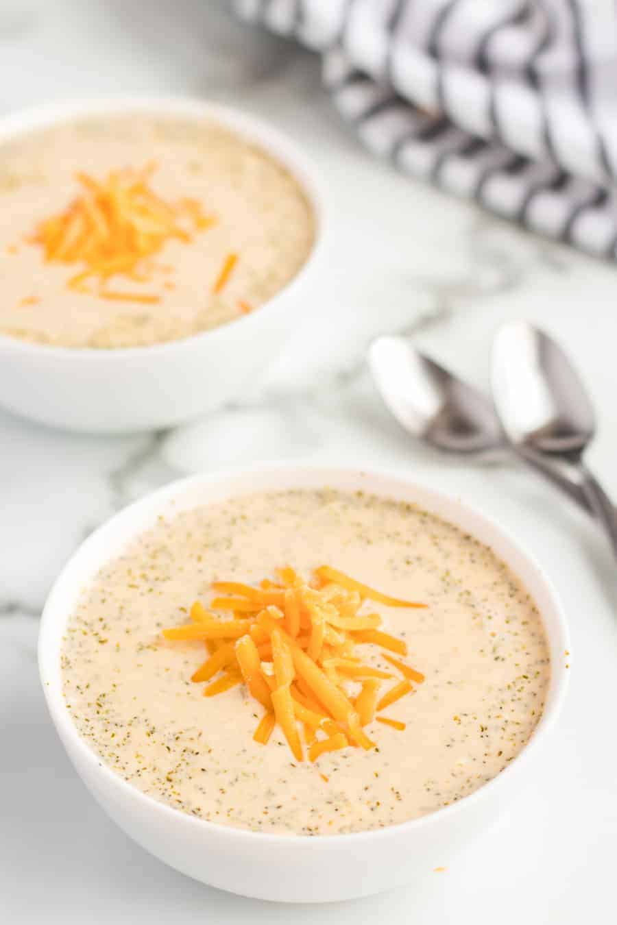 Two white bowls filled with creamy soup topped with shredded cheddar cheese sit on a marble surface, next to two spoons and a folded striped cloth in the background.