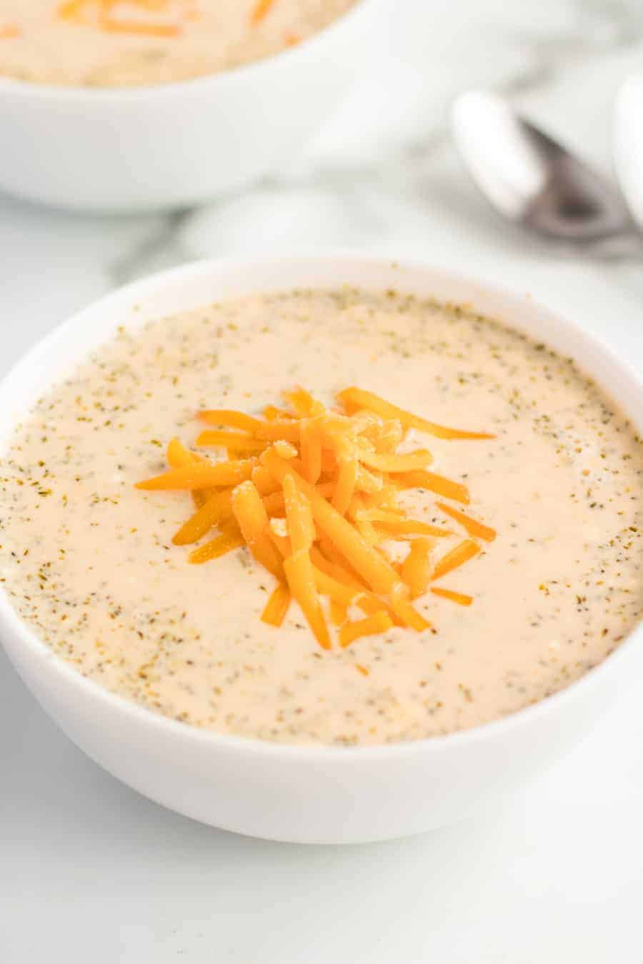 A white bowl filled with creamy crockpot broccoli and cheese soup, topped with shredded cheddar. A spoon and another bowl are visible in the blurred background.