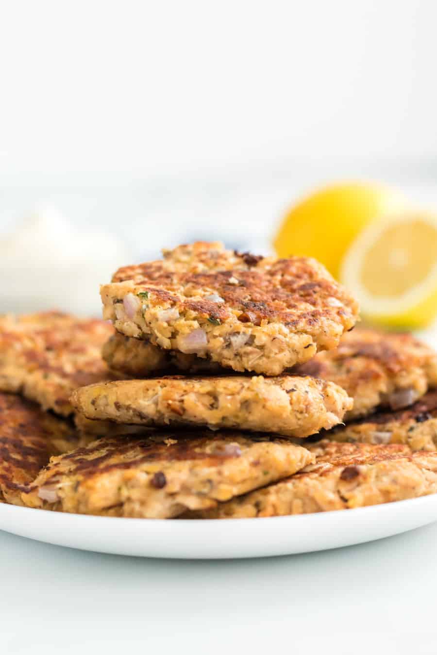 A plate of golden-brown salmon patties stacked on top of each other. Fresh lemon halves are blurred in the background, adding a hint of vibrant color. The patties appear crispy and well-cooked.