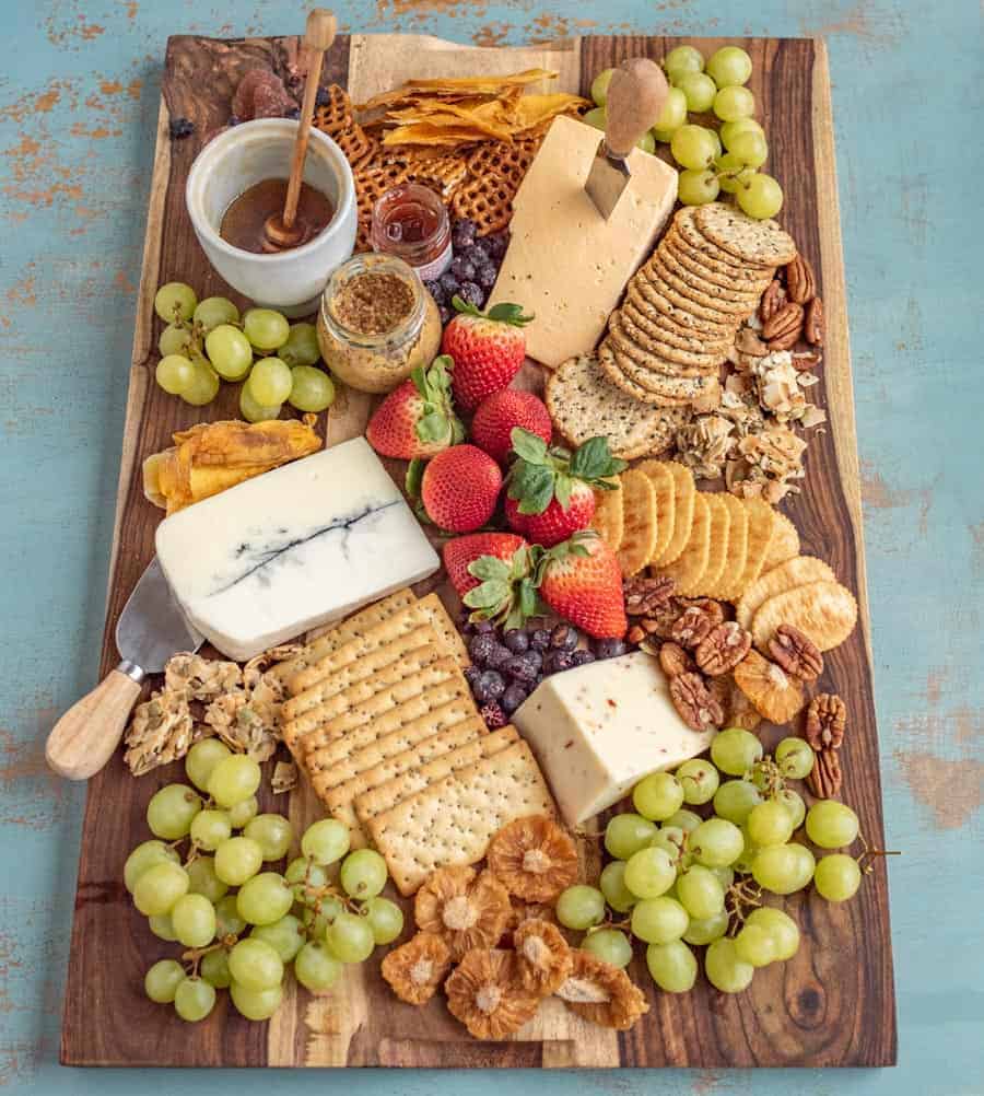 A cheese & charcuterie board on a wooden platter features assorted cheeses, crackers, green grapes, strawberries, nuts, dried fruits, a small jar of honey with a dipper, and mustard—all set atop a blue surface.