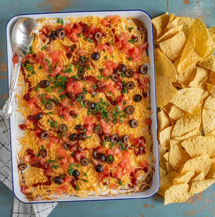 layered bean dip with cheddar cheese, tomatoes, black olives, and green onions on top with pile of tortilla chips beside it and a spoon in it.
