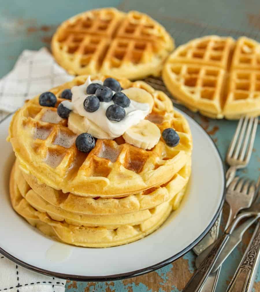 A four stack of Belgian Waffles on a white plate with bananas, blueberries, and whipped cream on top.