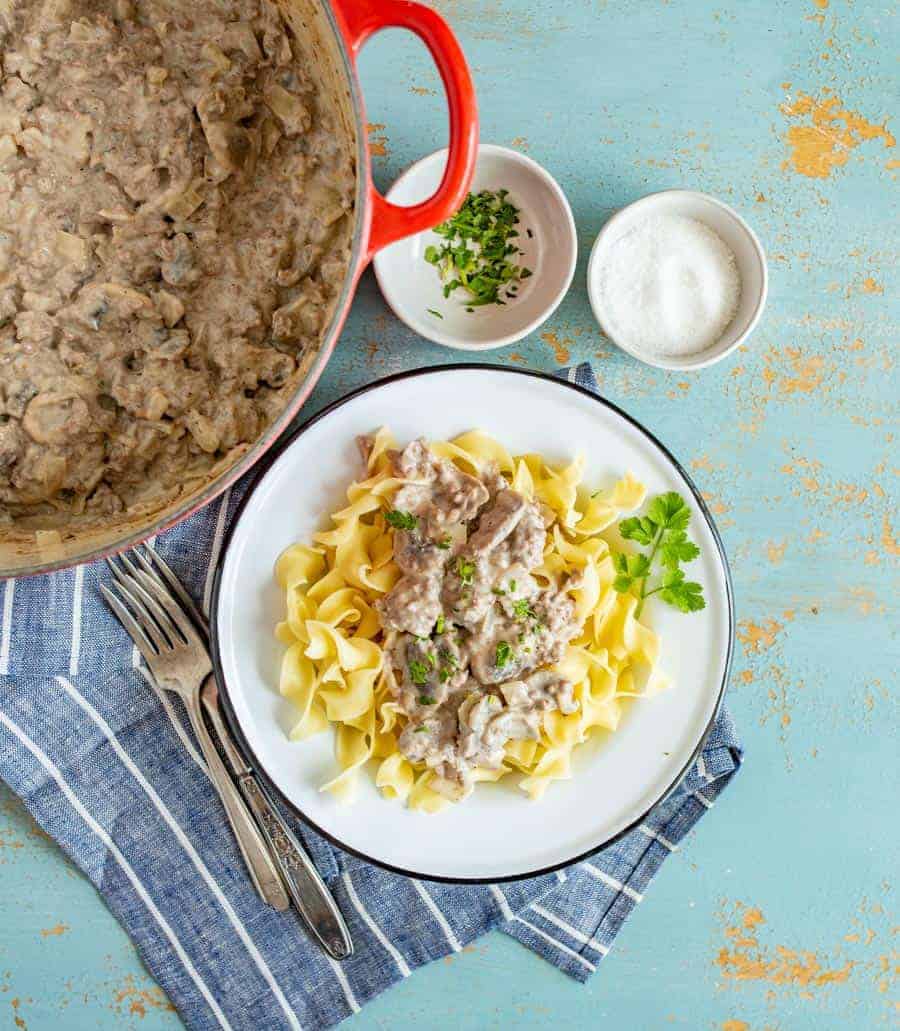 egg noddles on a white plate with ground beef and white sauce on top with green parsley and forks next to it with white bowls and red pan in background.