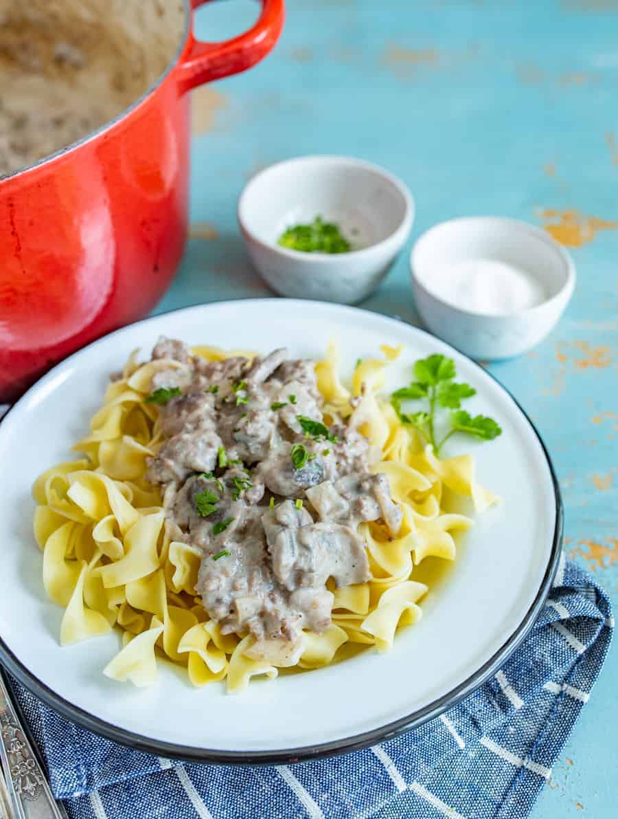 Homemade Ground beef stroganoff recipe showing egg noddles on a white plate with ground beef and white sauce on top with green parsley and white bowls and red pan in background.