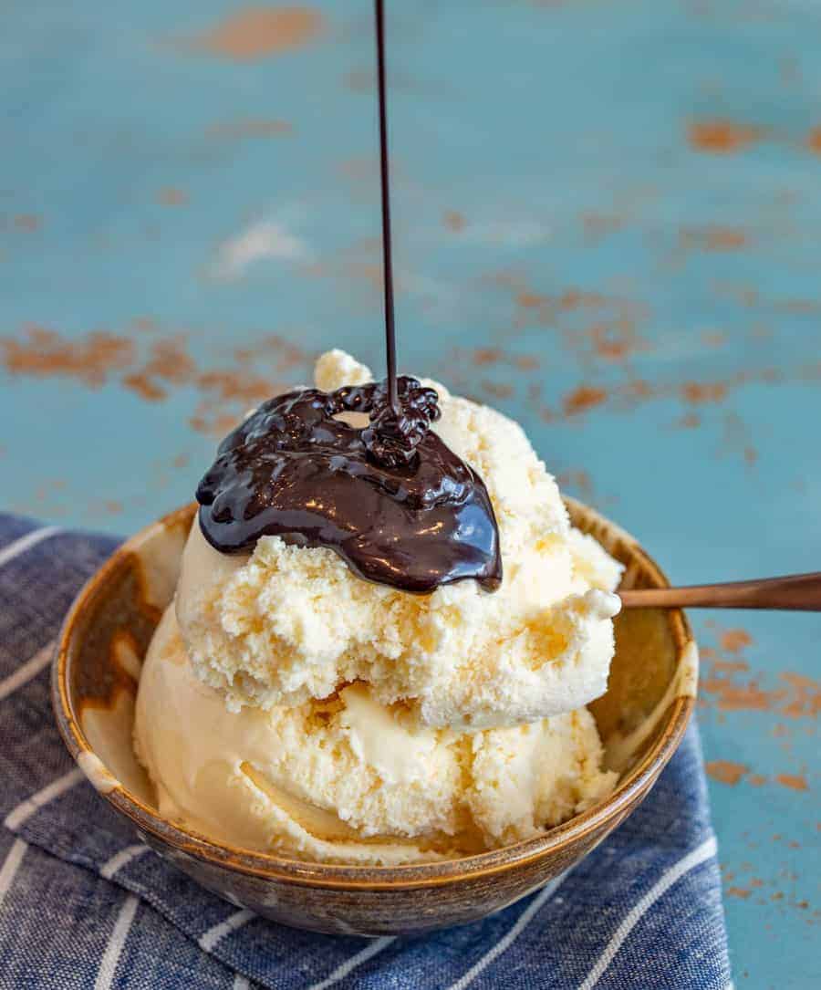 A bowl of vanilla ice cream with hot fudge sauce recipe poured over the top, sitting on a blue napkin with a spoon in the bowl against a rustic blue background.