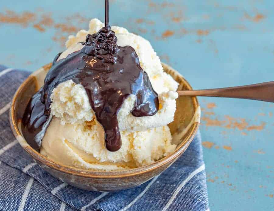 A bowl of vanilla ice cream is topped with thick chocolate sauce, made from a rich hot fudge sauce recipe, with a spoon resting in the bowl. The bowl sits on a blue and white striped cloth, on a light blue surface.