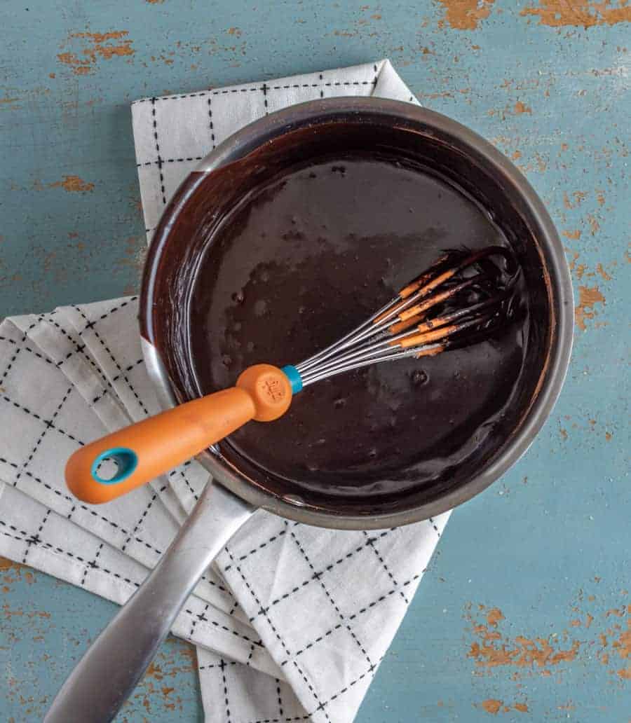 A saucepan filled with glossy chocolate sauce, perfect for your favorite hot fudge sauce recipe, and an orange-handled whisk rest on a white checkered cloth atop a distressed blue surface.