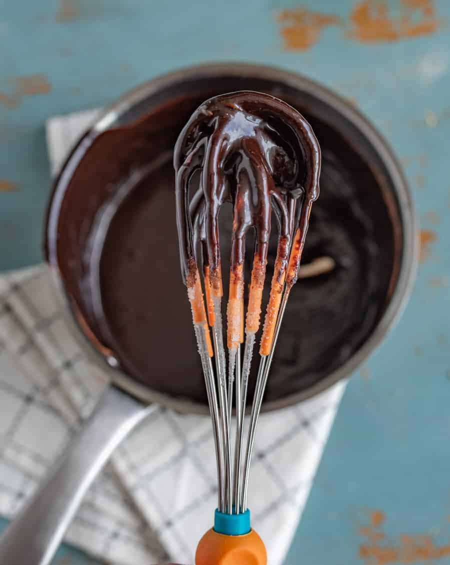 A whisk covered in glossy, melted chocolate is held above a saucepan filled with more chocolate, hinting at a rich hot fudge sauce recipe, with a white plaid kitchen towel underneath.