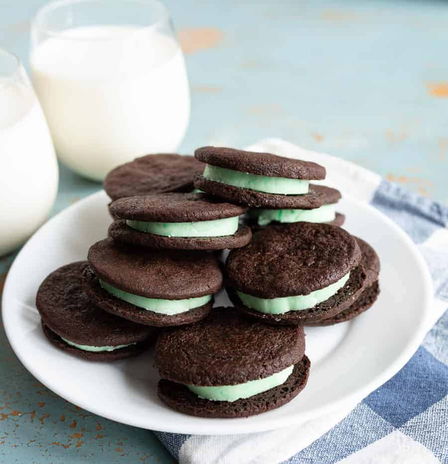 pile of homemade mint oreo cookies with dark chocolate outside and mint green inside stacked on a white plate on a blue and white towel on a blue background with two clear glasses of white milk beside them.