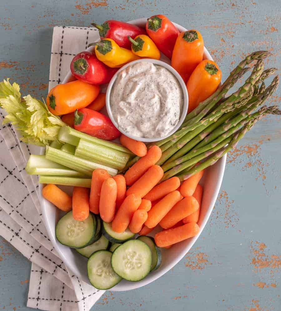white bowl of colorful vegetables with a bowl of creamy homemade ranch dressing dip on a black and white towel.