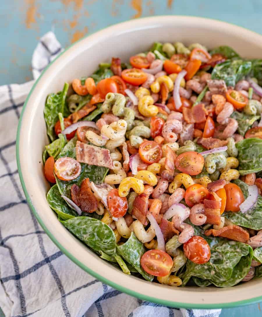 pasta salad with different colored vegetables in a big bowl with wooden spoons sitting on a blue and white towel.
