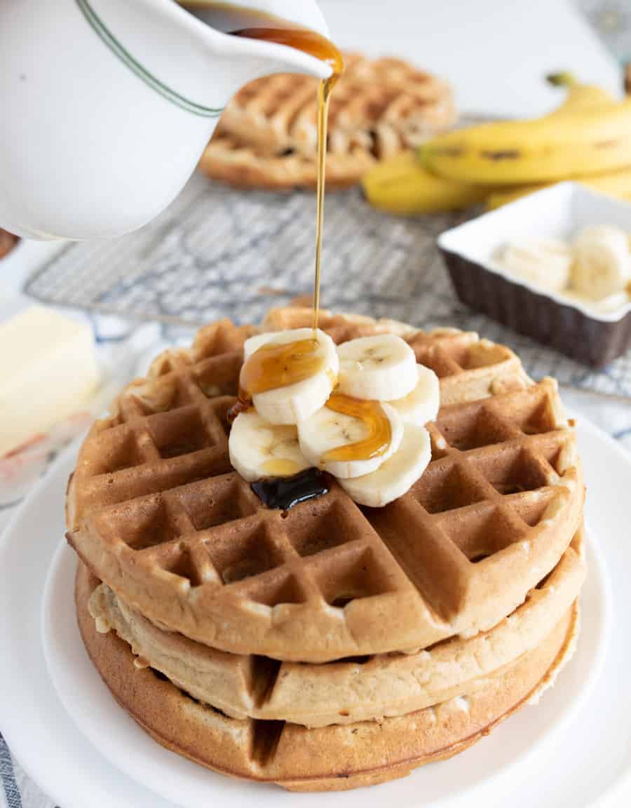 A stack of banana waffles topped with fresh banana slices is being drizzled with syrup from a white pitcher. In the background, ripe bananas accompany a dish of butter and another waffle resting on a cooling rack.
