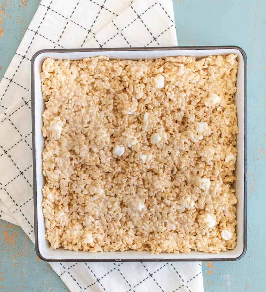 A square baking pan filled with a homemade rice krispie treats recipe sits on a white, checkered cloth over a blue surface. The treats appear lightly golden with visible marshmallow pieces mixed in.