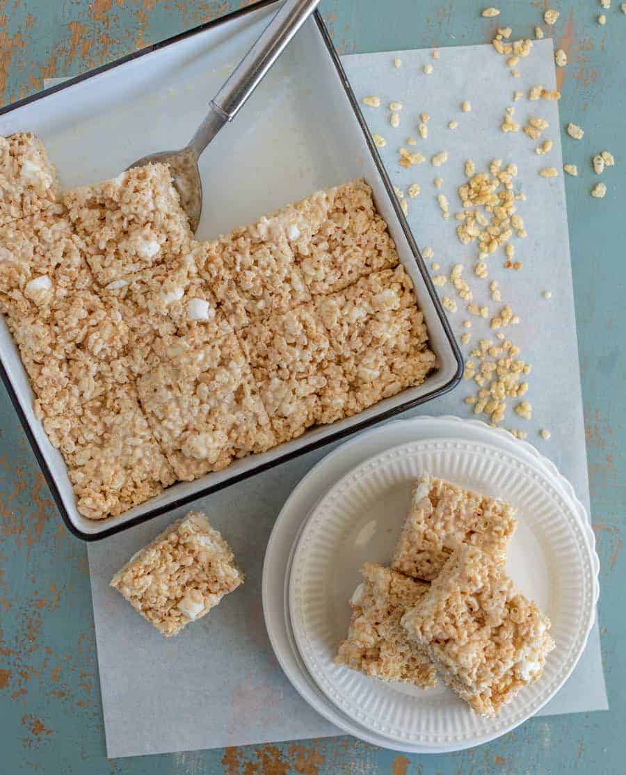 A pan of cut Rice Krispie treats recipe sits on a blue distressed surface, with a metal spatula in the pan. Three treats are stacked on white plates, and cereal pieces are scattered nearby.