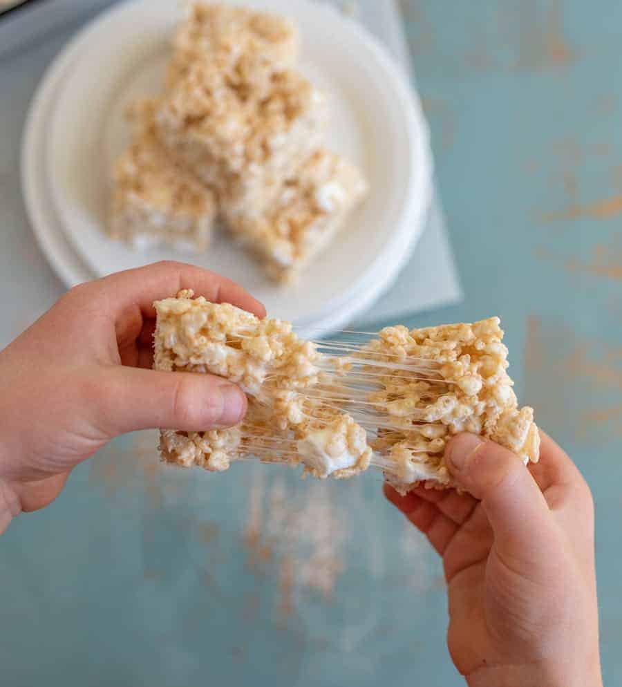 A person pulls apart a gooey rice krispie treat with both hands, stretching the marshmallow filling. A plate with more treats and a homemade rice krispie treats recipe sits on a blue table in the background.