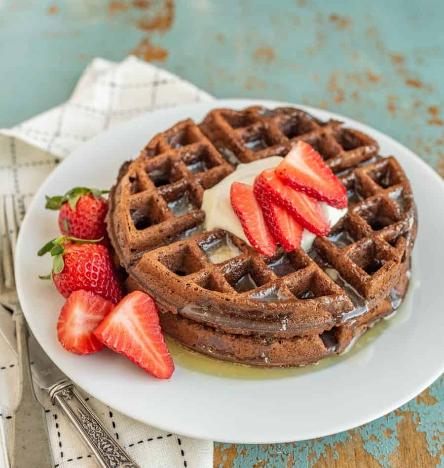 choclate waffle on a white plate with whole strawberries beside them and sliced strawberries, butter, and syrup on top.