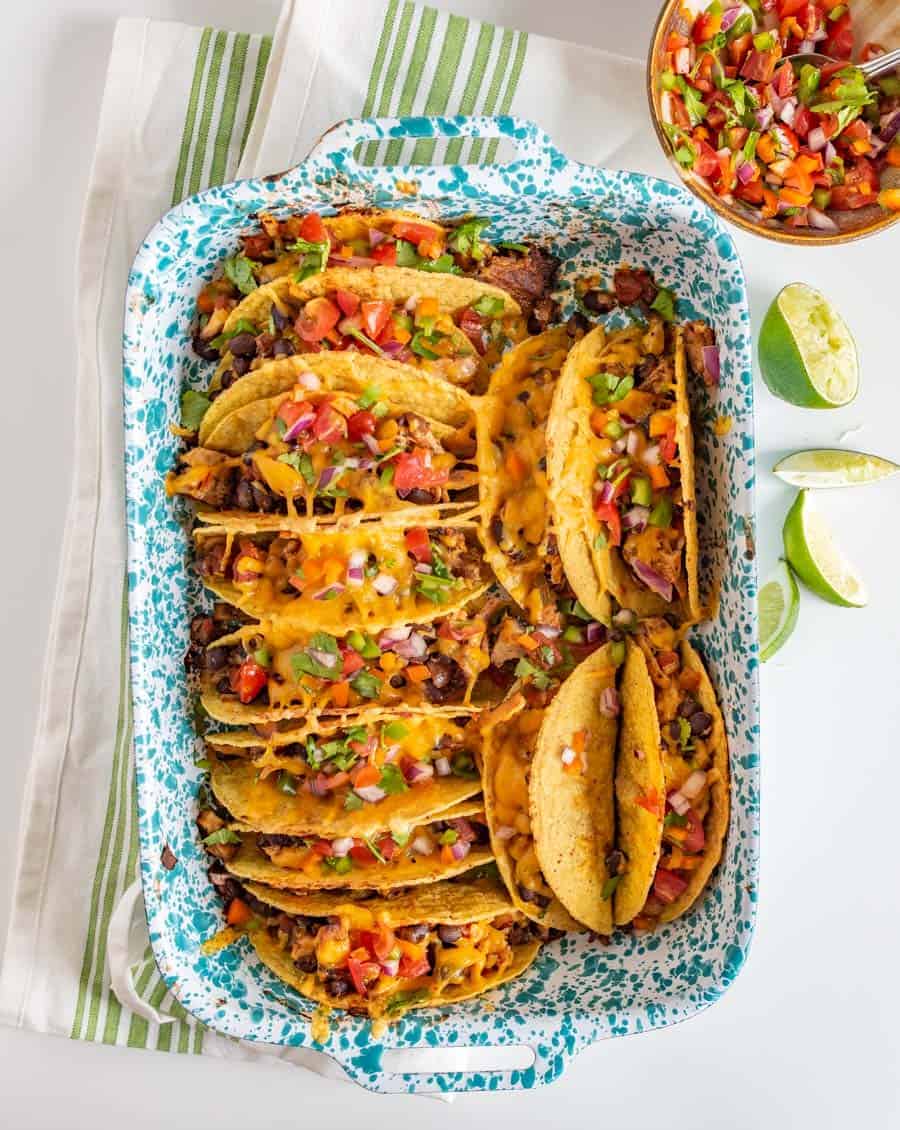 A baking dish holds several cheesy baked chicken tacos filled with meat, beans, and topped with diced tomatoes, onions, and herbs. Lime wedges and a bowl of salsa are on the side. The dish sits on a striped kitchen towel.