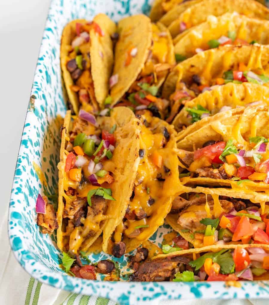 A baking dish filled with baked chicken tacos, topped with melted cheese, diced tomatoes, onions, and cilantro. The tacos are arranged upright and packed closely together, displaying vibrant ingredients.