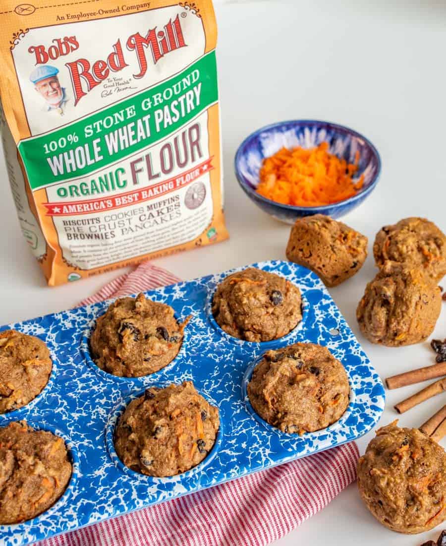 muffins in a blue and white muffin pan with small bowl of shredded carrot, cinnamon sticks, raisins, and a bag of flour on a red and white striped towel.