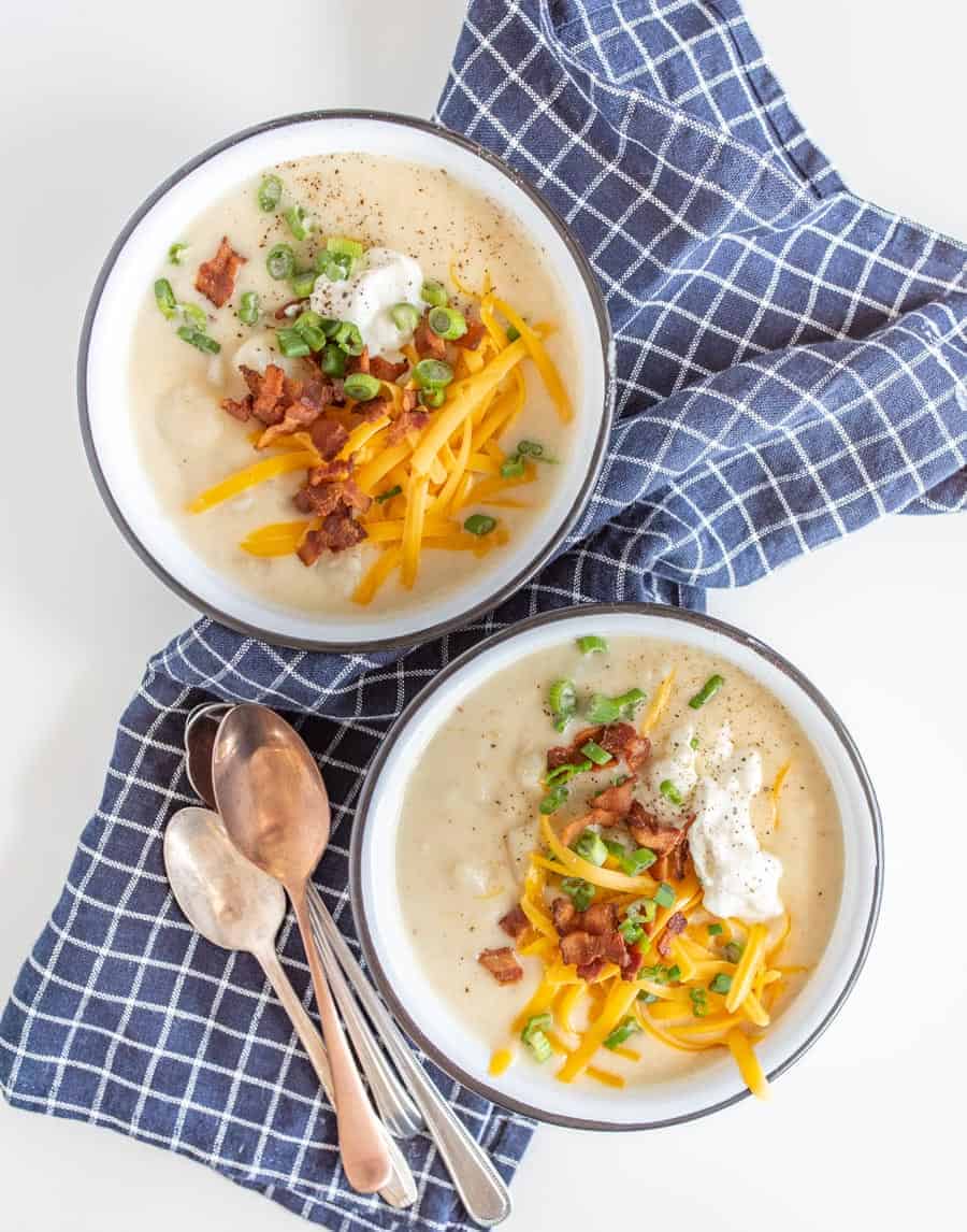 potato soup in white bowls with yellow cheddar cheese, sliced green onions, bacon bits, and sour cream on top sitting on a blue and white towel with spoons beside.