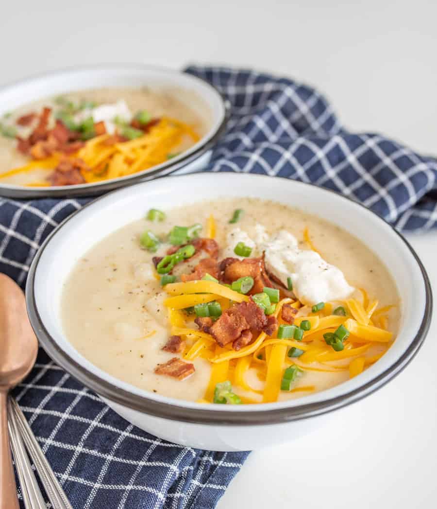 image for instant pot loaded baked potato soup showing potato soup in white bowls with yellow cheddar cheese, sliced green onions, bacon bits, and sour cream on top sitting on a blue and white towel with spoons beside.