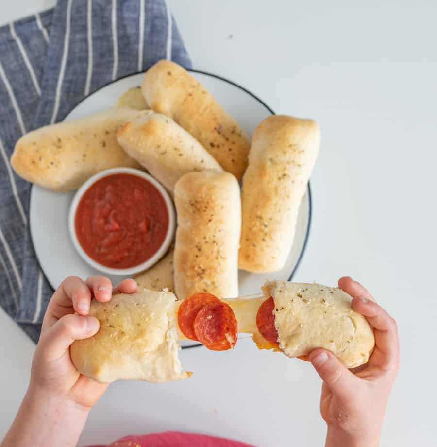breadsticks on a white plate with a small bowl of red marinara sauce on a blue and white striped towel with hand holding one bread stick with cheese and pepperoni coming out of it.