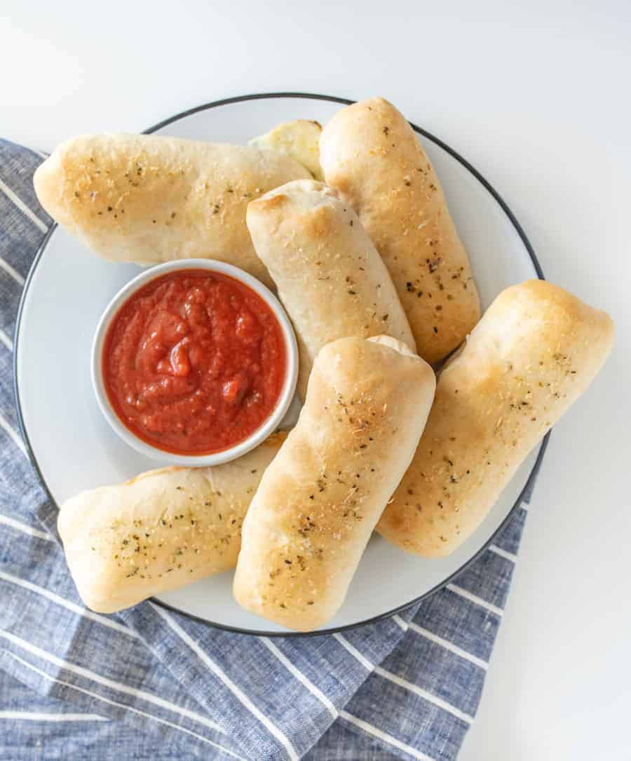 pepperoni pizza sticks on a white plate with a small bowl of red marinara sauce on a blue and white striped towel.