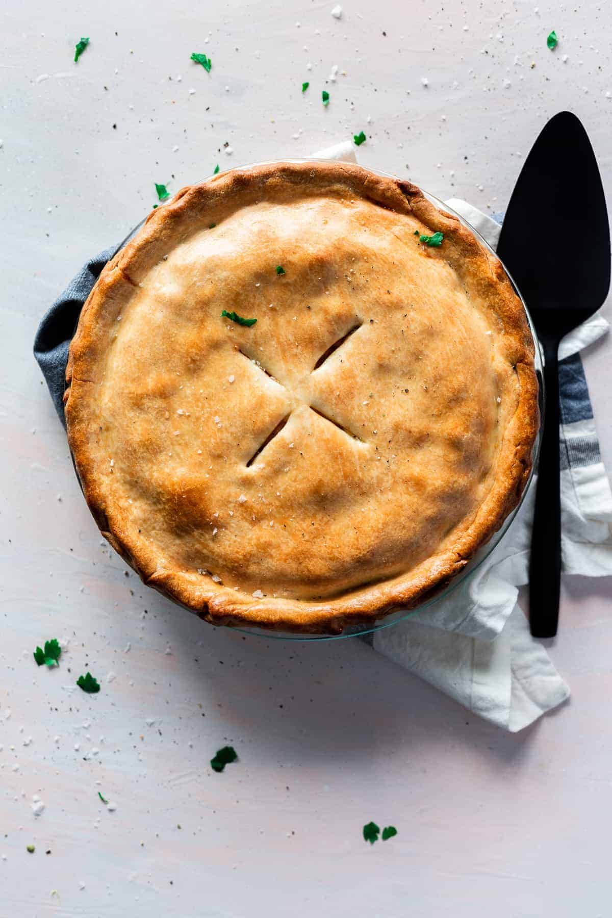 A golden-brown pie with a flaky crust, made from an easy chicken pot pie recipe, sits on a light surface next to a black pie server and white towel. Small green herb leaves are scattered around.