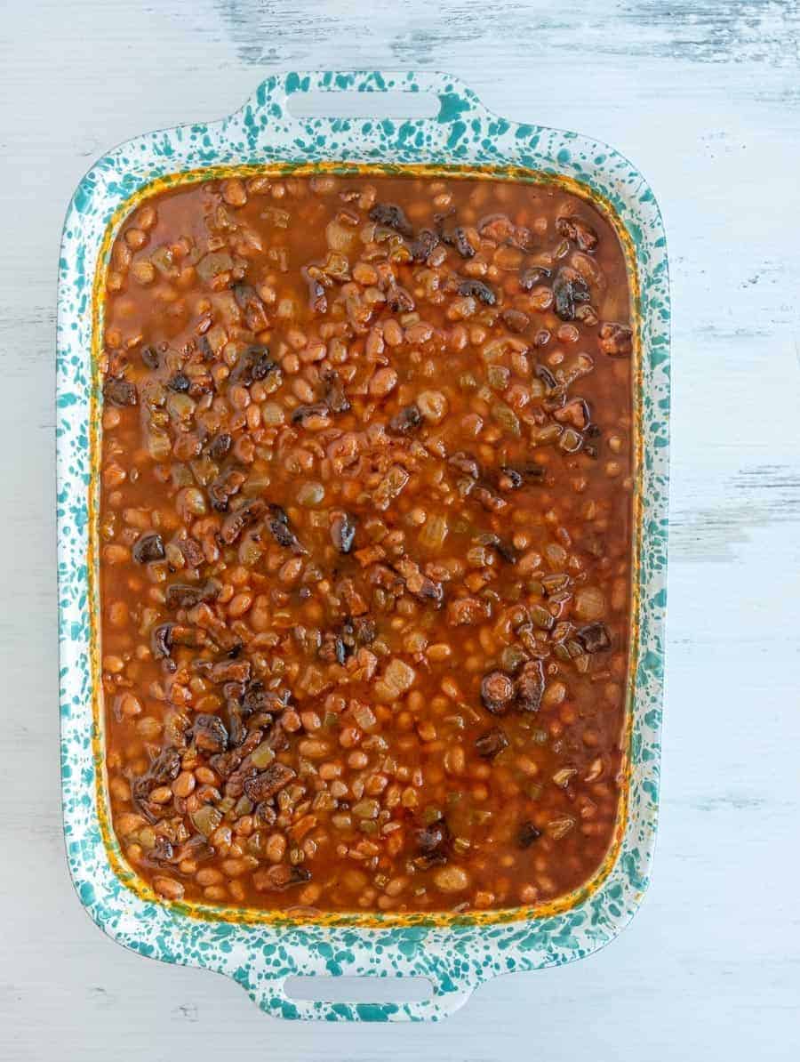 A blue speckled baking dish brimming with savory baked beans, crowned with chunks of crispy bacon. The beans are enveloped in a rich, dark sauce, creating a mouthwatering display as it rests on a white textured surface.