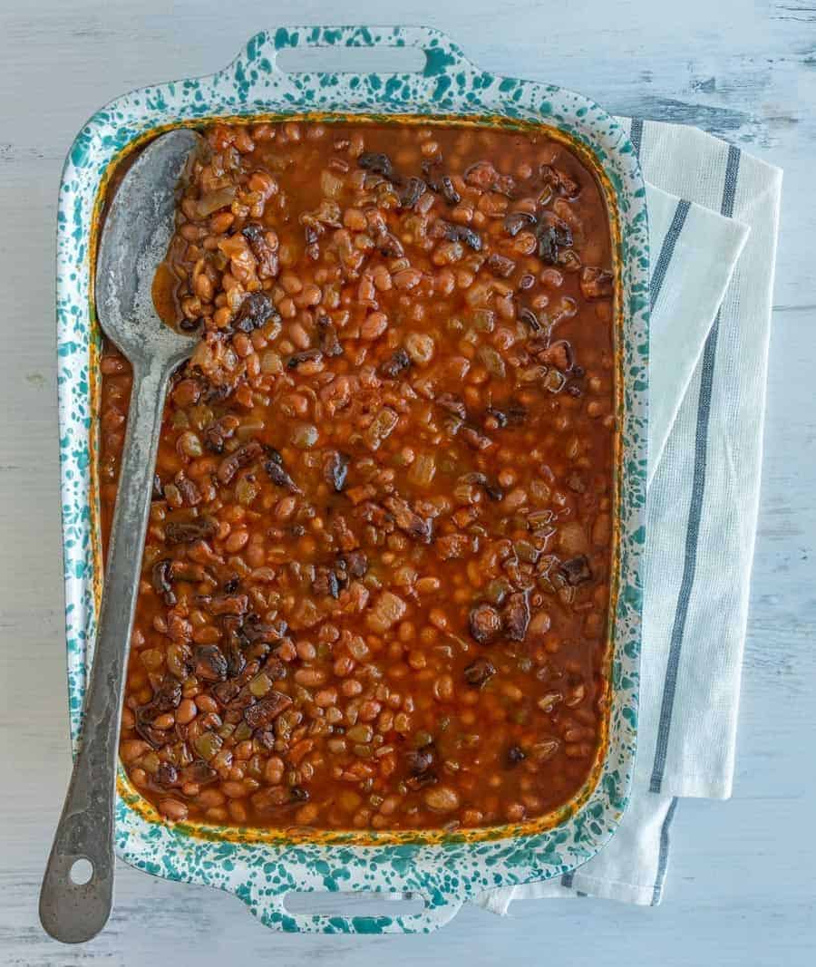A baking dish brimming with savory baked beans in a rich, dark sauce sits invitingly. A large serving spoon rests on top, ready to serve. The dish is elegantly placed on a white wooden surface, complemented by a white and gray striped cloth underneath.