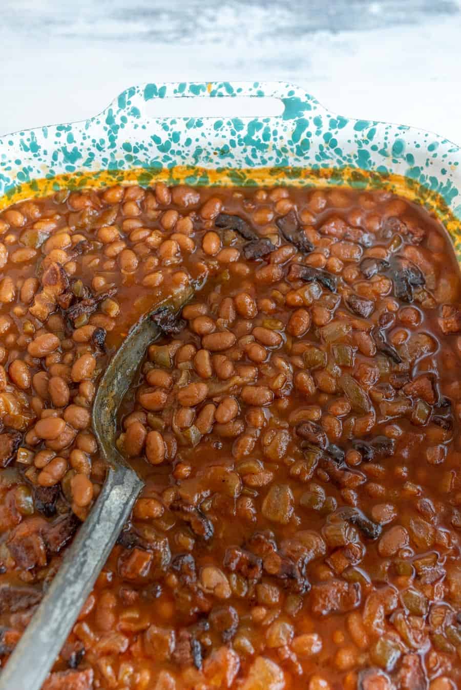 A close-up of baked beans recipe in a blue-speckled rectangular dish invites you to explore the delicious baked beans recipe. The beans swim in a thick sauce, and a metal spoon rests inside, scooping from one side. The surface shows some charred bits, suggesting they are freshly baked.