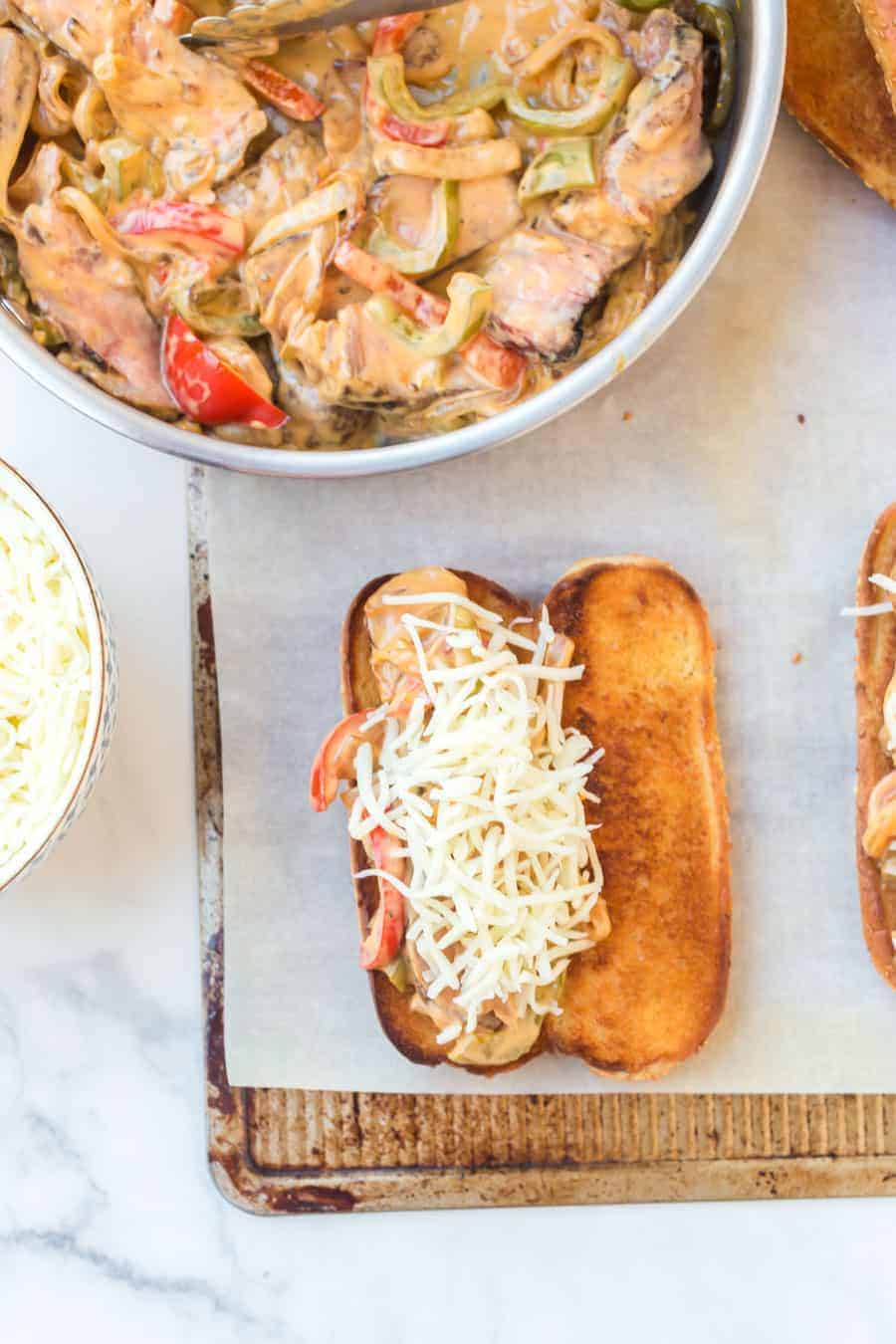 sesame bun grilled and open with shredded cheese. metal pan filled with steak, onions, peppers and melted cheese in background.