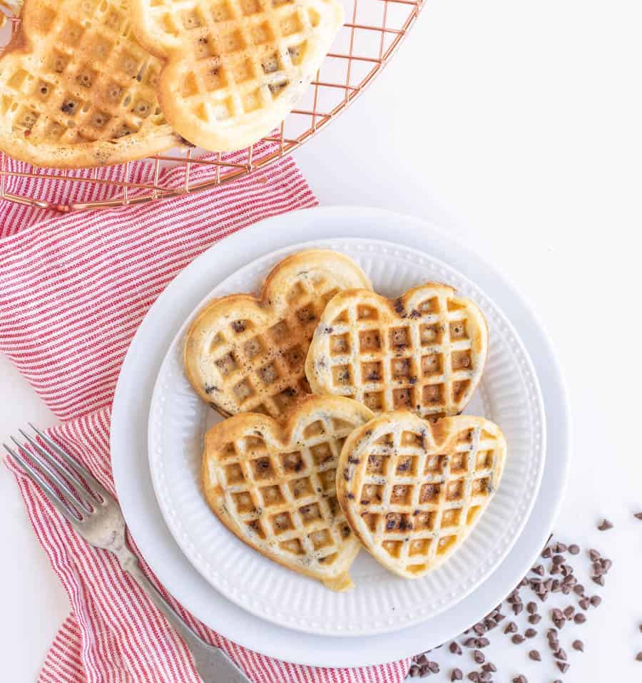 Heart-shaped waffles with chocolate chips are stacked on a white plate, ready to be paired with ice cream, all on a red-striped cloth. A fork rests nearby while more waffles tempt from a wire rack in the background, with small chocolate chips playfully scattered around.