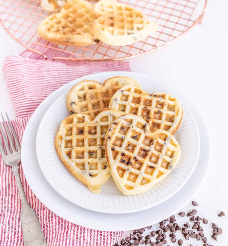 Heart-shaped chocolate chip waffles stacked on a pristine white plate. A red-striped cloth and fork rest to the left, while additional chocolatey delights cool on a wire rack in the background, surrounded by scattered chips for an extra touch of sweetness.
