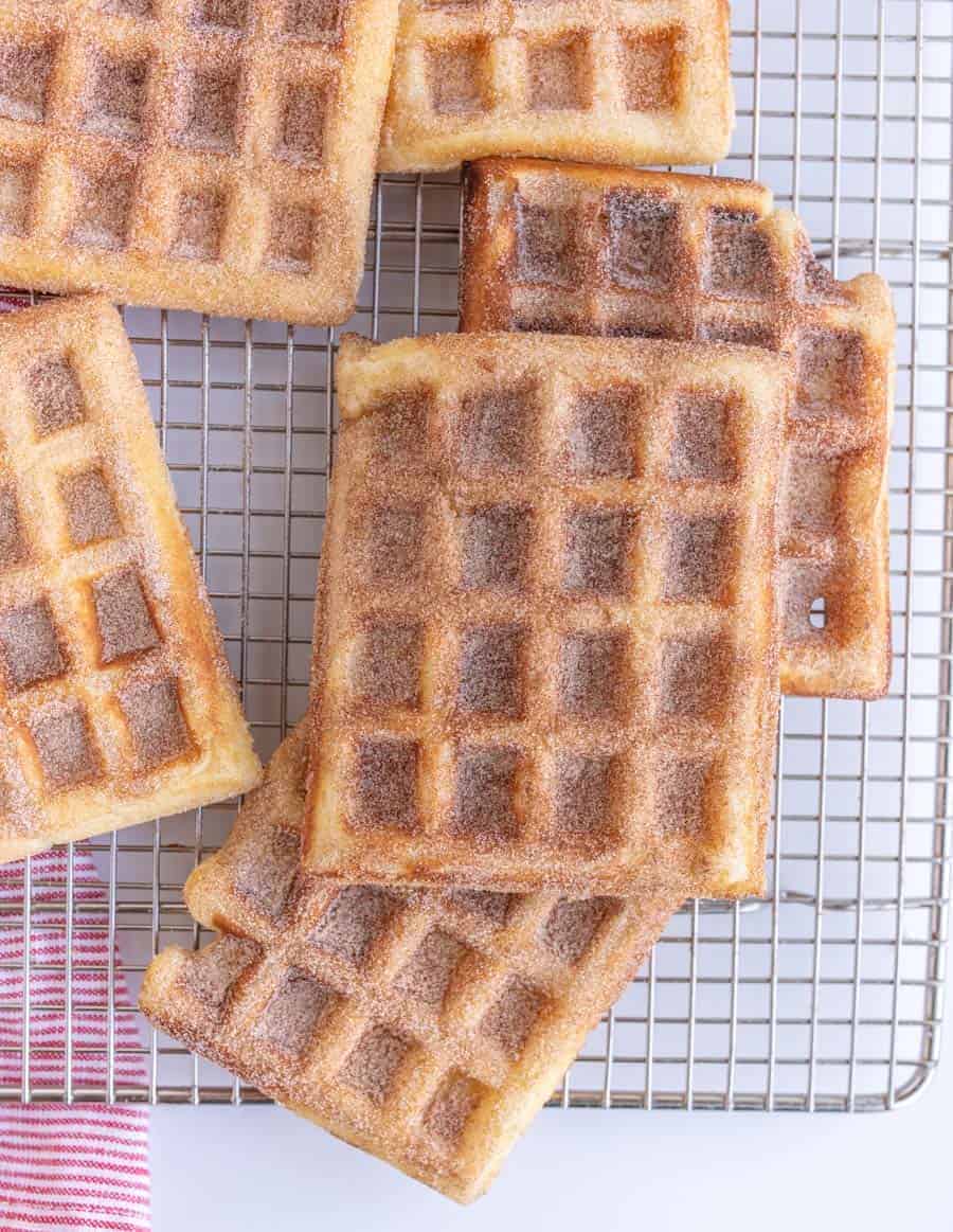 Several golden-brown waffle slices are arranged on a cooling rack beside a scoop of velvety ice cream. A red and white striped cloth is partially visible in the bottom left corner. The waffles have a sugary coating, giving them a crisp texture.