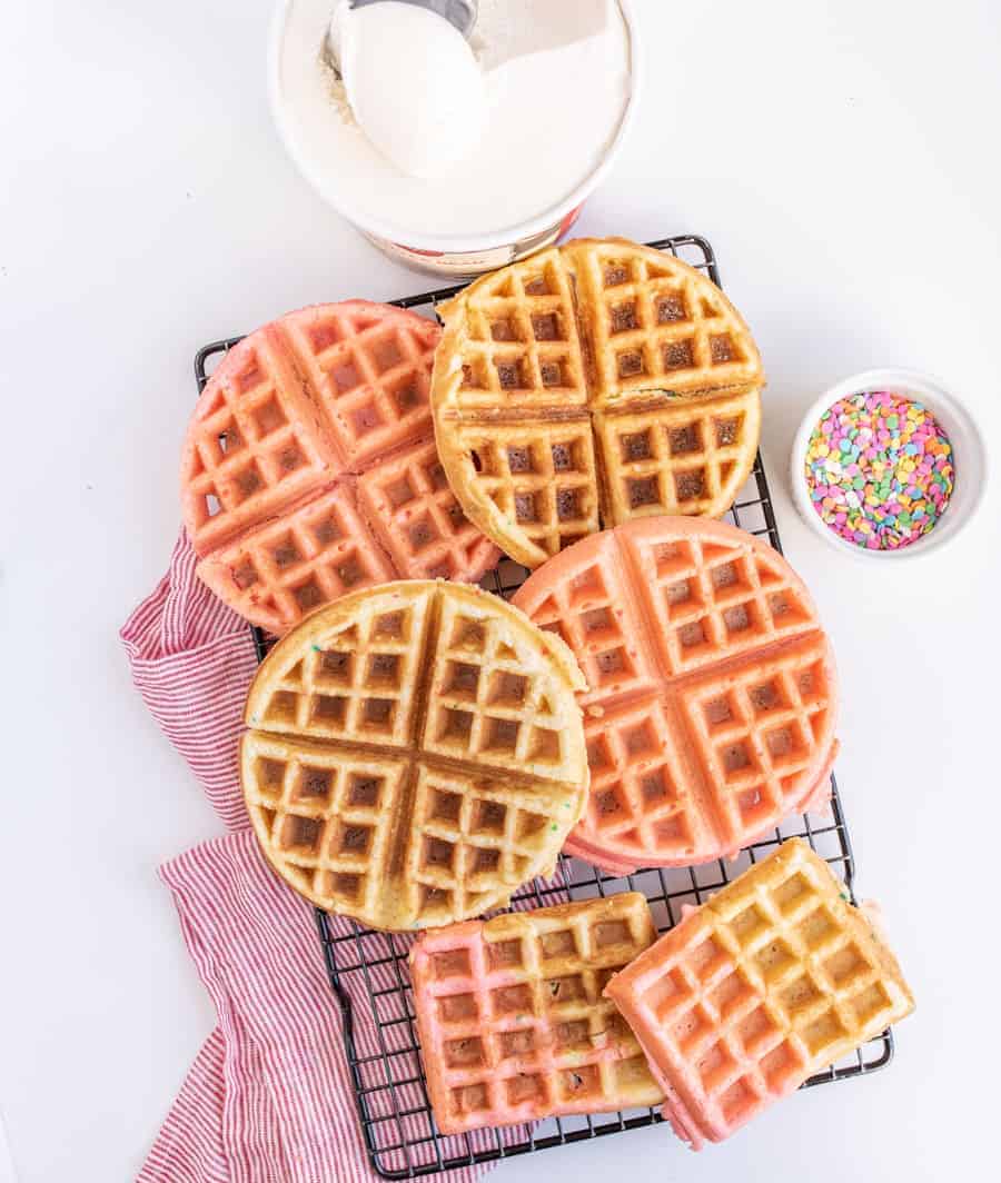 A cooling rack holds round pink and golden brown waffles on top of a red-striped cloth. Nearby are a tub of ice cream with a scoop and a bowl of colorful sprinkles, all set against a white background.
