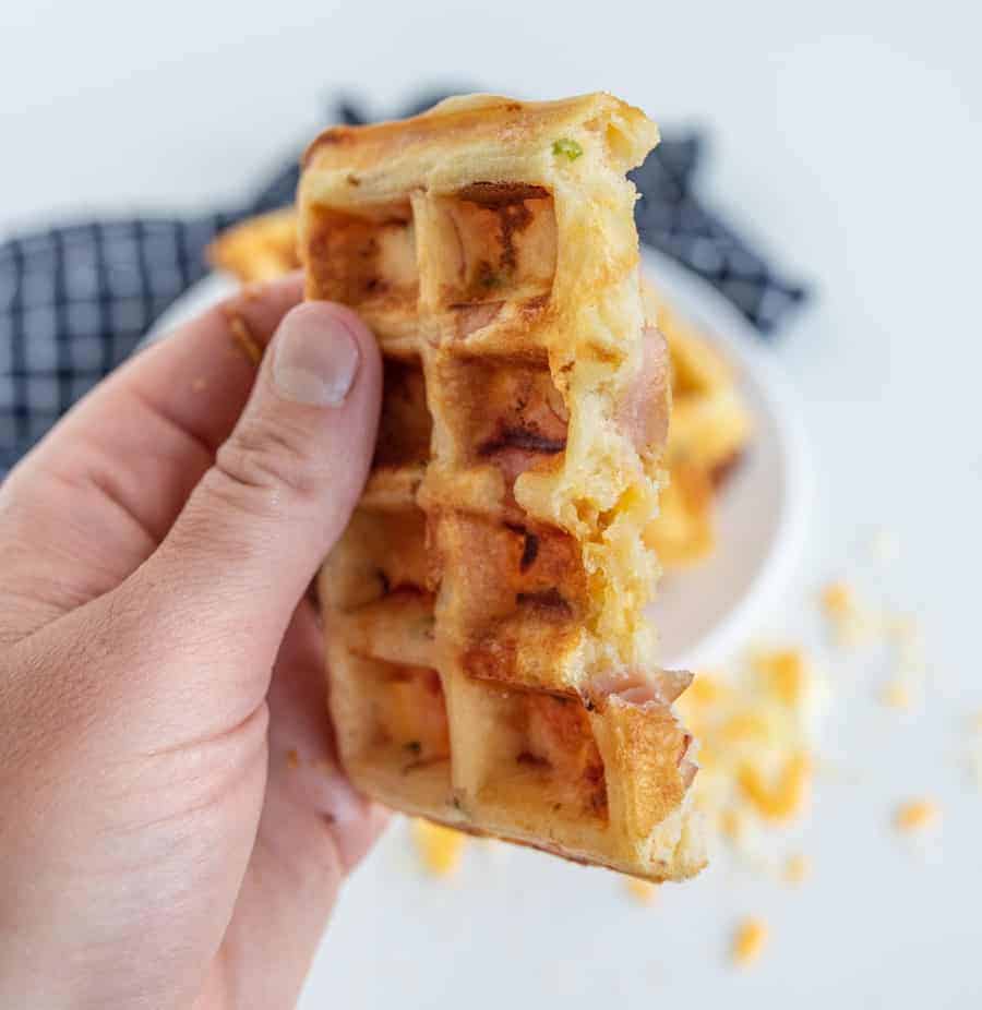 A hand holds a partially eaten waffle, laden with cheese and bacon chunks. Crumbs scatter below, while the background blurs into a black-and-white checkered cloth. Imagine adding a scoop of creamy ice cream for an indulgent twist on this savory delight.