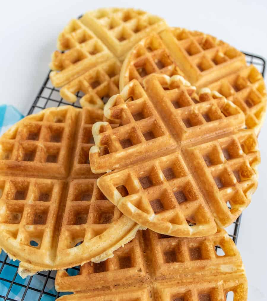 Golden, round waffles stacked on a cooling rack. The waffles have a crisp texture with grid patterns on top, and they are set against a light background.