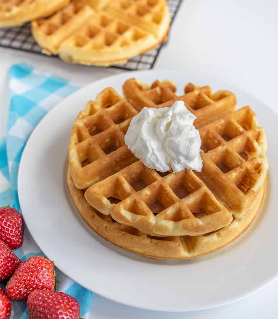 A stack of two golden waffles topped with a dollop of whipped cream sits on a white plate. Fresh strawberries are to the side, with more waffles cooling on a wire rack in the background. A blue and white checkered cloth rests under the plate.