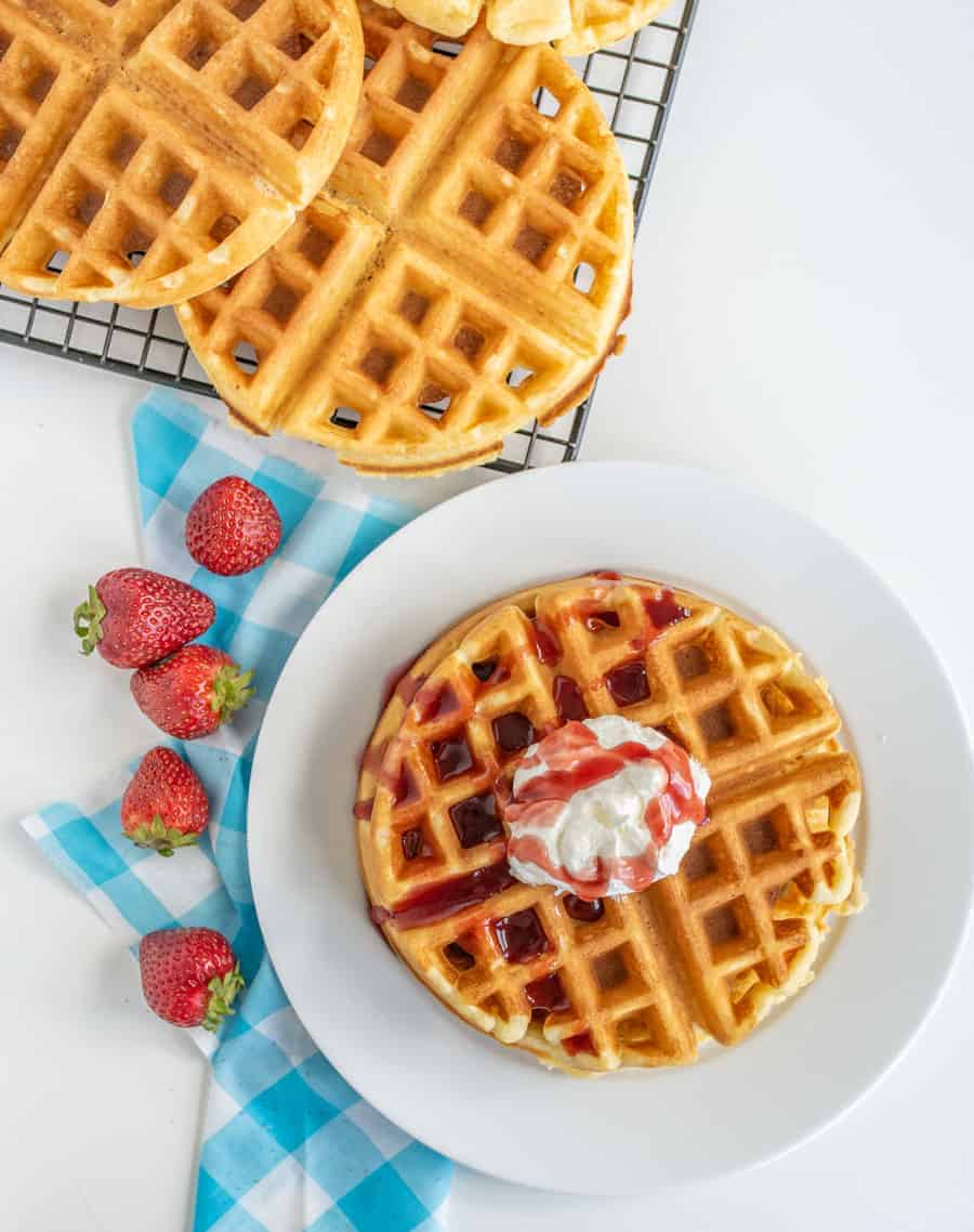 A plate with a fluffy waffle from my favorite fluffy waffle recipe is topped with strawberry syrup and whipped cream, sitting on a blue checkered napkin. Three fresh strawberries are beside it. Behind the plate, more waffles rest on a cooling rack.