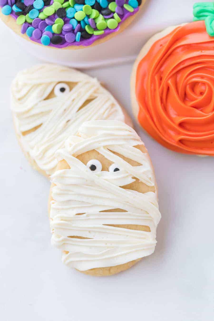 sugar cookies decorated with halloween theme white frosting to look like a mummy with candy eyes and colored sprinkles in purple black green and blue colors.