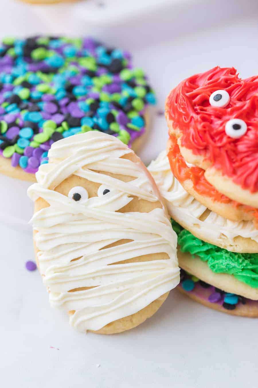 sugar cookies decorated with halloween theme white frosting to look like a mummy with candy eyes and colored sprinkles in purple black green and blue colors.