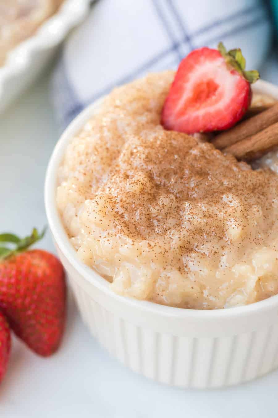 A white bowl filled with the best rice pudding recipe, topped with cinnamon, a sliced strawberry, and cinnamon sticks, with whole strawberries beside the bowl.