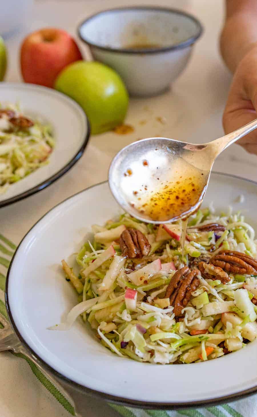 upclose image of apple cabbage salad topped with pecans with a dressing being spooned on top.