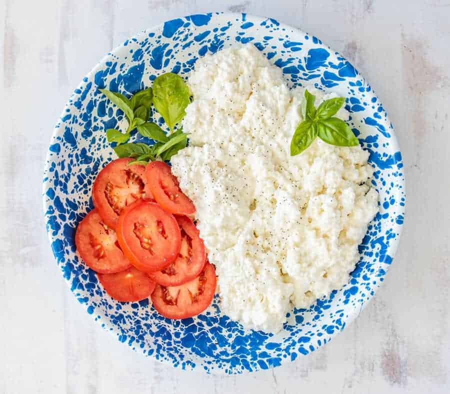 top view of fresh cottage cheese on a blue speckled plate with fresh basil and sliced tomatoes