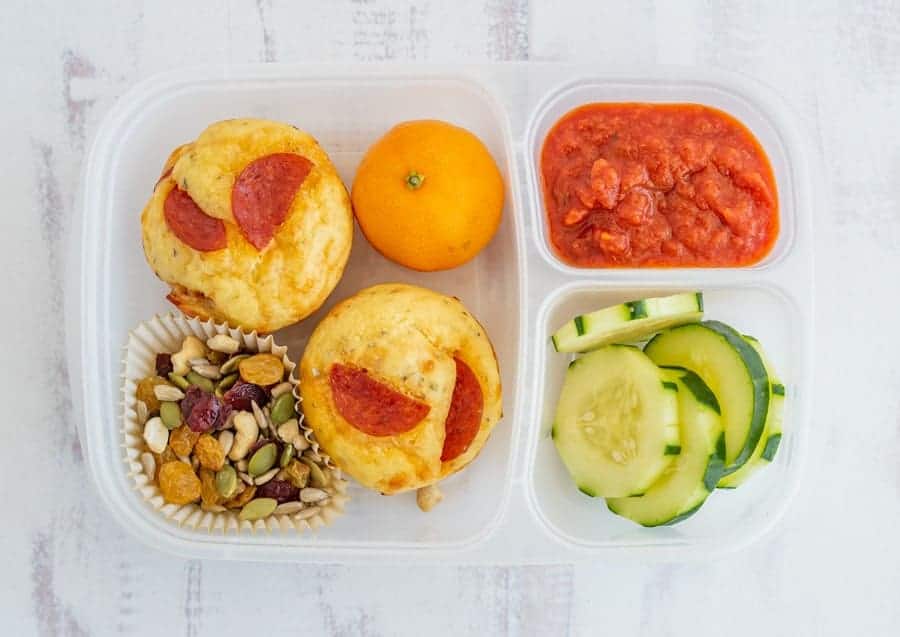 to-go lunch box with pizza bites, an orange, trail mix, cucumbers, and marinara sauce.