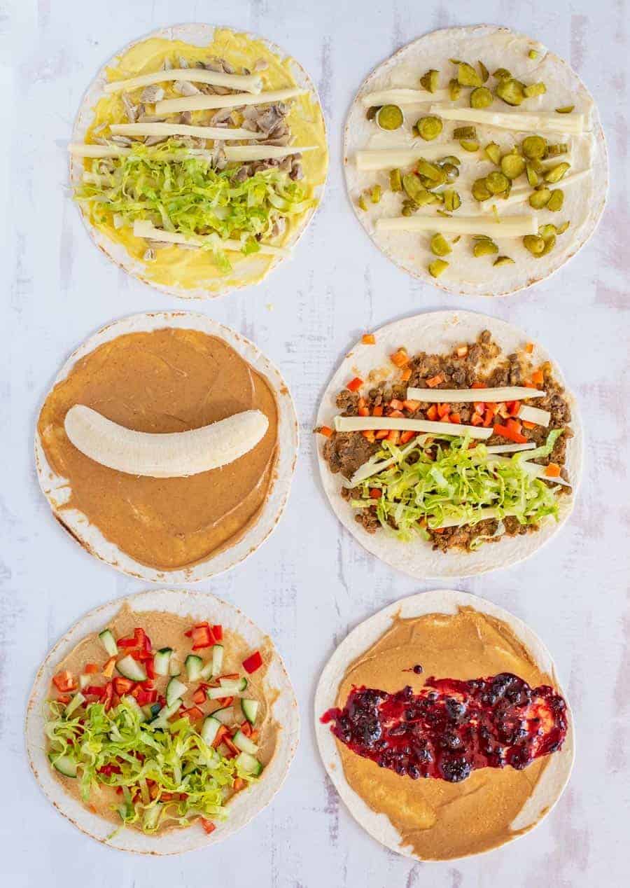 Six tortillas on a light surface, each topped with ingredients like lettuce, cheese, pickles, banana, peanut butter, veggies, ground meat, and berry jam—perfect inspiration for easy pinwheel recipes. Arranged in two vertical rows of three.