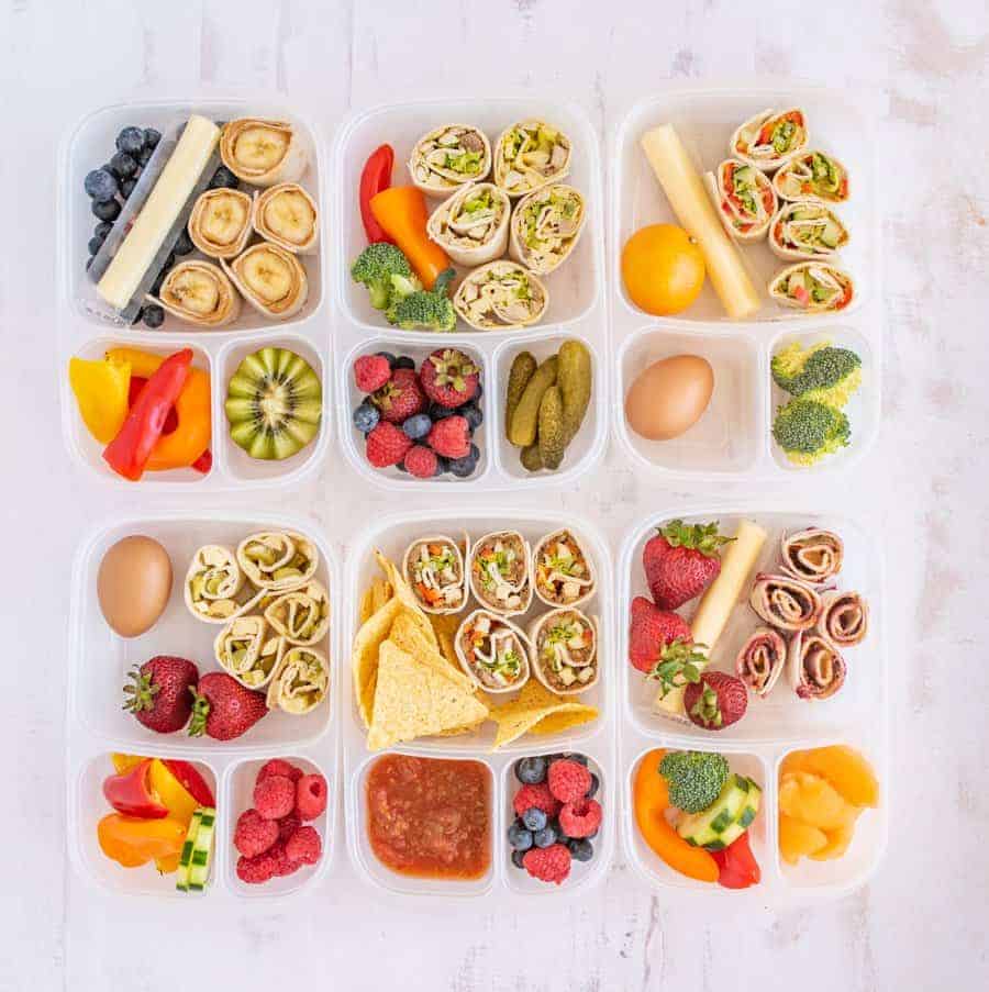 Lunchboxes filled with a variety of snacks, including easy pinwheel recipes, fruits (berries, kiwi, strawberries), vegetables (broccoli, peppers, cucumber), boiled eggs, cheese sticks, chips with salsa, and pickles.